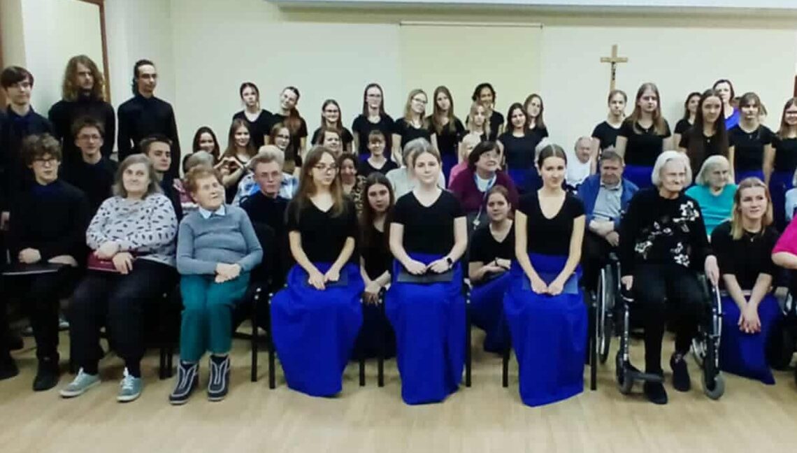 Grupowe zdjęcie kilkudziesięciu osób w różnym wieku, ustawionych w kilku rzędach w sali. Większość osób ubrana jest na czarno; część kobiet ma długie, intensywnie niebieskie spódnice. W pierwszym rzędzie siedzą osoby starsze, w tym osoby poruszające się na wózkach inwalidzkich. W tle jasna ściana z krzyżem, drewniana podłoga. Scena sugeruje wspólne wydarzenie artystyczne lub koncert o charakterze integracyjnym.