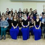 Grupowe zdjęcie kilkudziesięciu osób w różnym wieku, ustawionych w kilku rzędach w sali. Większość osób ubrana jest na czarno; część kobiet ma długie, intensywnie niebieskie spódnice. W pierwszym rzędzie siedzą osoby starsze, w tym osoby poruszające się na wózkach inwalidzkich. W tle jasna ściana z krzyżem, drewniana podłoga. Scena sugeruje wspólne wydarzenie artystyczne lub koncert o charakterze integracyjnym.