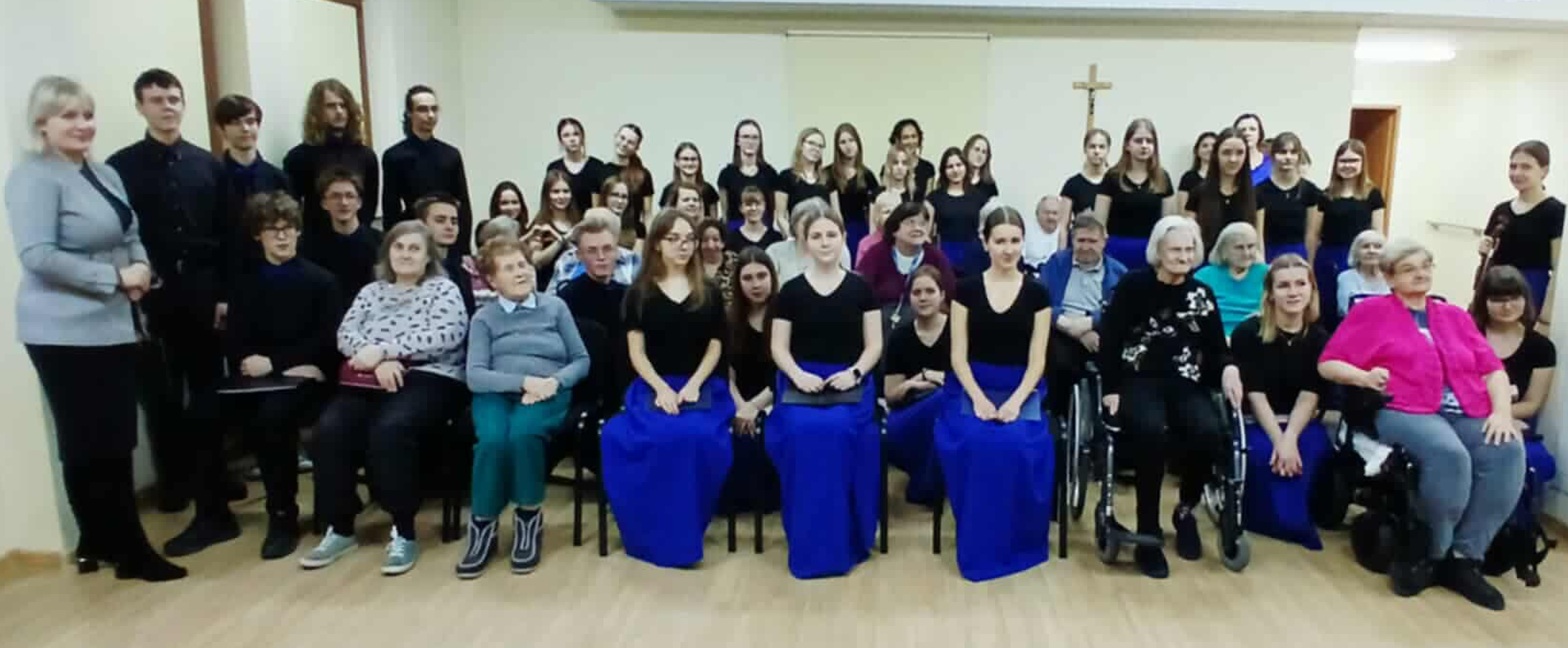 Grupowe zdjęcie kilkudziesięciu osób w różnym wieku, ustawionych w kilku rzędach w sali. Większość osób ubrana jest na czarno; część kobiet ma długie, intensywnie niebieskie spódnice. W pierwszym rzędzie siedzą osoby starsze, w tym osoby poruszające się na wózkach inwalidzkich. W tle jasna ściana z krzyżem, drewniana podłoga. Scena sugeruje wspólne wydarzenie artystyczne lub koncert o charakterze integracyjnym.