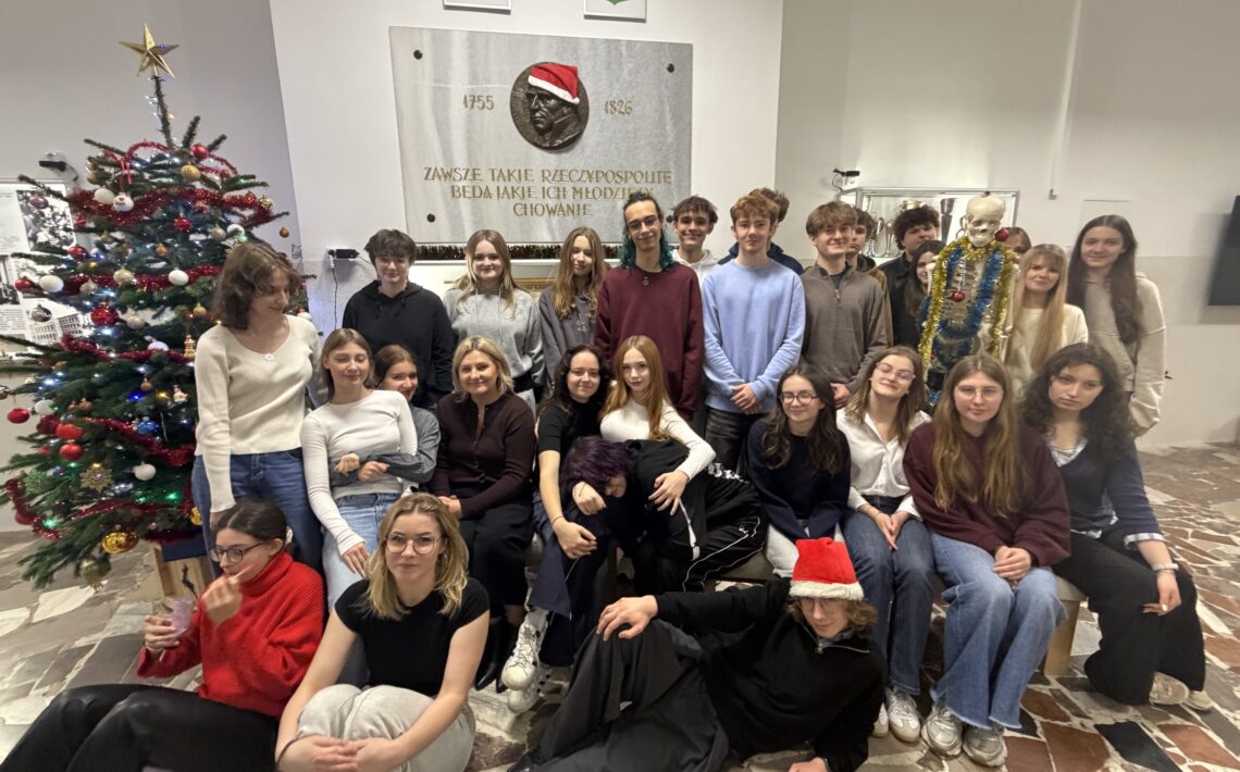 Zdjęcie przedstawia dużą grupę młodych osób — prawdopodobnie uczniów szkoły średniej — pozujących wspólnie w przestrzeni szkolnej podczas świątecznego spotkania. Łącznie na zdjęciu znajduje się około 25 osób, w większości nastolatków, a także jedna dorosła kobieta (nauczycielka). Scena jest jasna i ciepła, oddająca atmosferę radości i integracji. Tło i otoczenie Główna ściana za grupą Zajmuje ją duża, jasna płyta pamiątkowa z brązowym medalionem — płaskorzeźbą przedstawiającą męską głowę w profilu. Medalion ma nałożoną czapkę Mikołaja: czerwoną, z białym futerkiem i pomponem. Po obu stronach medalionu widnieją lata „1755” i „1826”. Pod portretem wypisany jest cytat w języku polskim: „Zawsze takie Rzeczypospolite będą, jakie ich młodzieży chowanie.” Litery są złote i eleganckie. Nad płytą wiszą dwa herby: po lewej herb Polski (biały orzeł w koronie na czerwonym tle), po prawej herb lokalny (biały motyw heraldyczny na czerwonym tle). Prawa strona zdjęcia Stoi tam szkielet anatomiczny w pełnym rozmiarze człowieka, ustawiony pionowo. Szkielet jest udekorowany na święta: owinięty złotą i niebieską lametą, na szyi ma bombki, wygląda żartobliwie i przyjaźnie. Lewa strona zdjęcia Znajduje się tam duża choinka bożonarodzeniowa, sięgająca prawie do sufitu. Ubrana jest w czerwone, białe i złote bombki, białe łańcuchy oraz zestaw światełek. Na czubku choinki świeci złota gwiazda. Pod drzewkiem widać stojak i kilka ozdób. Podłoga Jest z jasnych i ciemnych kamiennych płytek ułożonych nieregularnie. Grupa osób – rozmieszczenie i wygląd Grupa jest ustawiona w trzech planach: siedzący i leżący z przodu, siedzący pośrodku, stojący z tyłu. Przód — osoby najbliżej obiektywu Lewy dolny róg Dziewczyna w czerwonym, miękkim swetrze; okulary; ciemne spodnie. Siedzi na podłodze, trzyma w dłoni różowy woreczek lub ozdobę i patrzy w dół. Obok niej — dziewczyna w czarnej koszulce i jasnych dresowych spodniach, siedzi prosto, patrzy w stronę aparatu. Centralnie z przodu Leży chłopak w czarnym stroju, w czapce Mikołaja. Jest oparty na zgiętym łokciu, z rozluźnioną, żartobliwą pozą. Środkowy rząd — osoby siedzące Po lewej siedzi nauczycielka/dorosła kobieta w bordowym swetrze i z blond włosami związanymi z tyłu. Wokół niej siedzą dziewczyny w jasnych i ciemnych swetrach, większość z długimi rozpuszczonymi włosami. Część osób obejmuje się wzajemnie, niektóre trzymają ręce na kolanach. Tylny rząd — osoby stojące To największa grupa — kilkanaście osób. Ubrania utrzymane są w spokojnych kolorach: czernie, szarości, granaty, bordo. Wszyscy uśmiechają się lub patrzą neutralnie w stronę aparatu. Po prawej stronie, tuż obok szkieletu, stoi znacznie wyższy chłopak w błękitnym swetrze. Po lewej — kilka osób stoi częściowo zasłoniętych przez choinkę. Atmosfera zdjęcia Zdjęcie jest wyraźnie wykonane z okazji Świąt Bożego Narodzenia — świadczy o tym choinka, dodatki świąteczne oraz czapki Mikołaja. Oświetlenie jest jasne, typowo szkolne, neutralne, bez efektów specjalnych. Grupa wygląda na zgraną, wyluzowaną, uśmiechniętą. Atmosfera jest ciepła i integrująca. Obecność świątecznie udekorowanego szkieletu dodaje zdjęciu humoru.
