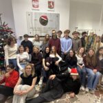 Zdjęcie przedstawia dużą grupę młodych osób — prawdopodobnie uczniów szkoły średniej — pozujących wspólnie w przestrzeni szkolnej podczas świątecznego spotkania. Łącznie na zdjęciu znajduje się około 25 osób, w większości nastolatków, a także jedna dorosła kobieta (nauczycielka). Scena jest jasna i ciepła, oddająca atmosferę radości i integracji. Tło i otoczenie Główna ściana za grupą Zajmuje ją duża, jasna płyta pamiątkowa z brązowym medalionem — płaskorzeźbą przedstawiającą męską głowę w profilu. Medalion ma nałożoną czapkę Mikołaja: czerwoną, z białym futerkiem i pomponem. Po obu stronach medalionu widnieją lata „1755” i „1826”. Pod portretem wypisany jest cytat w języku polskim: „Zawsze takie Rzeczypospolite będą, jakie ich młodzieży chowanie.” Litery są złote i eleganckie. Nad płytą wiszą dwa herby: po lewej herb Polski (biały orzeł w koronie na czerwonym tle), po prawej herb lokalny (biały motyw heraldyczny na czerwonym tle). Prawa strona zdjęcia Stoi tam szkielet anatomiczny w pełnym rozmiarze człowieka, ustawiony pionowo. Szkielet jest udekorowany na święta: owinięty złotą i niebieską lametą, na szyi ma bombki, wygląda żartobliwie i przyjaźnie. Lewa strona zdjęcia Znajduje się tam duża choinka bożonarodzeniowa, sięgająca prawie do sufitu. Ubrana jest w czerwone, białe i złote bombki, białe łańcuchy oraz zestaw światełek. Na czubku choinki świeci złota gwiazda. Pod drzewkiem widać stojak i kilka ozdób. Podłoga Jest z jasnych i ciemnych kamiennych płytek ułożonych nieregularnie. Grupa osób – rozmieszczenie i wygląd Grupa jest ustawiona w trzech planach: siedzący i leżący z przodu, siedzący pośrodku, stojący z tyłu. Przód — osoby najbliżej obiektywu Lewy dolny róg Dziewczyna w czerwonym, miękkim swetrze; okulary; ciemne spodnie. Siedzi na podłodze, trzyma w dłoni różowy woreczek lub ozdobę i patrzy w dół. Obok niej — dziewczyna w czarnej koszulce i jasnych dresowych spodniach, siedzi prosto, patrzy w stronę aparatu. Centralnie z przodu Leży chłopak w czarnym stroju, w czapce Mikołaja. Jest oparty na zgiętym łokciu, z rozluźnioną, żartobliwą pozą. Środkowy rząd — osoby siedzące Po lewej siedzi nauczycielka/dorosła kobieta w bordowym swetrze i z blond włosami związanymi z tyłu. Wokół niej siedzą dziewczyny w jasnych i ciemnych swetrach, większość z długimi rozpuszczonymi włosami. Część osób obejmuje się wzajemnie, niektóre trzymają ręce na kolanach. Tylny rząd — osoby stojące To największa grupa — kilkanaście osób. Ubrania utrzymane są w spokojnych kolorach: czernie, szarości, granaty, bordo. Wszyscy uśmiechają się lub patrzą neutralnie w stronę aparatu. Po prawej stronie, tuż obok szkieletu, stoi znacznie wyższy chłopak w błękitnym swetrze. Po lewej — kilka osób stoi częściowo zasłoniętych przez choinkę. Atmosfera zdjęcia Zdjęcie jest wyraźnie wykonane z okazji Świąt Bożego Narodzenia — świadczy o tym choinka, dodatki świąteczne oraz czapki Mikołaja. Oświetlenie jest jasne, typowo szkolne, neutralne, bez efektów specjalnych. Grupa wygląda na zgraną, wyluzowaną, uśmiechniętą. Atmosfera jest ciepła i integrująca. Obecność świątecznie udekorowanego szkieletu dodaje zdjęciu humoru.