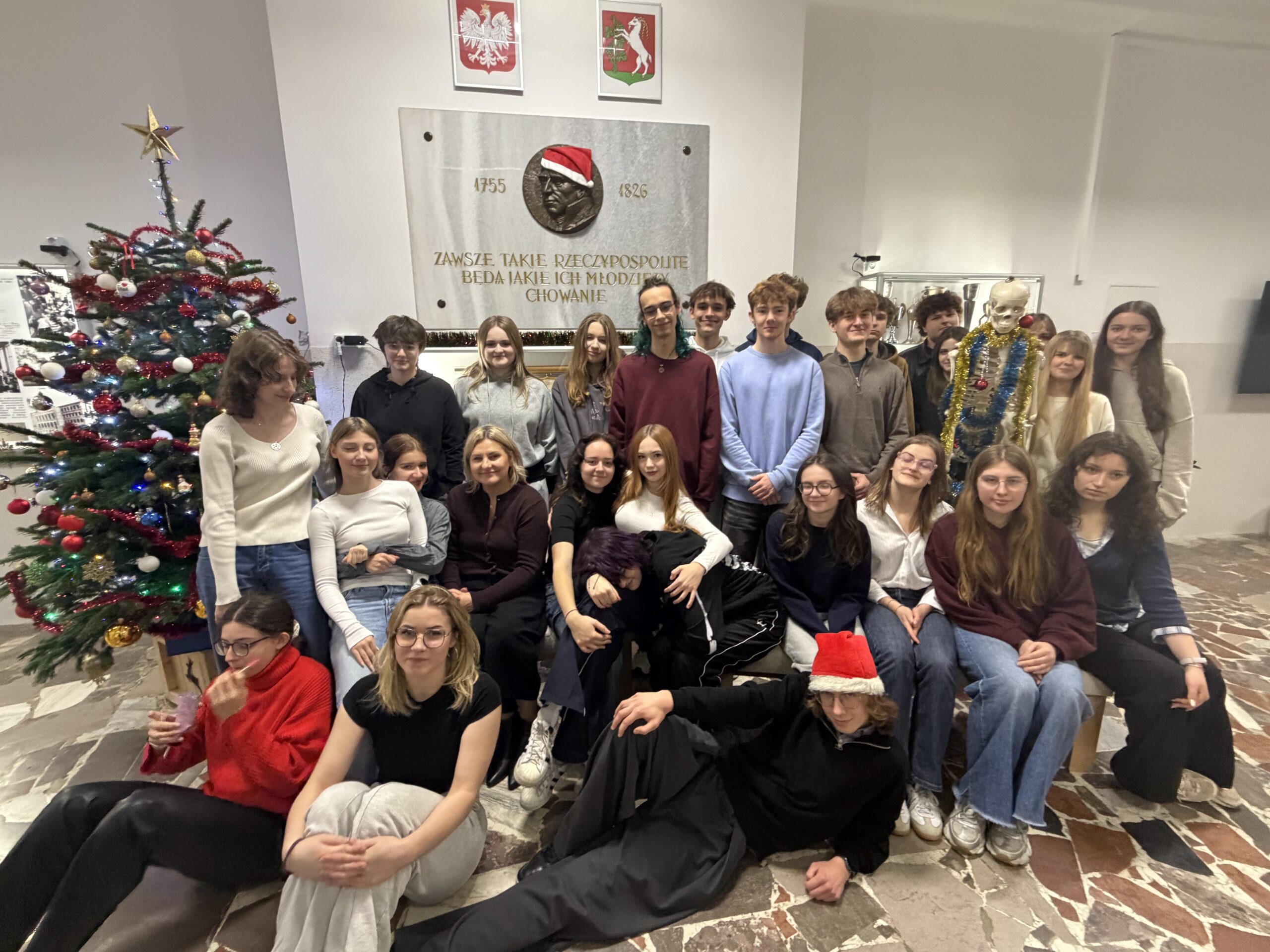 Zdjęcie przedstawia dużą grupę młodych osób — prawdopodobnie uczniów szkoły średniej — pozujących wspólnie w przestrzeni szkolnej podczas świątecznego spotkania. Łącznie na zdjęciu znajduje się około 25 osób, w większości nastolatków, a także jedna dorosła kobieta (nauczycielka). Scena jest jasna i ciepła, oddająca atmosferę radości i integracji. Tło i otoczenie Główna ściana za grupą Zajmuje ją duża, jasna płyta pamiątkowa z brązowym medalionem — płaskorzeźbą przedstawiającą męską głowę w profilu. Medalion ma nałożoną czapkę Mikołaja: czerwoną, z białym futerkiem i pomponem. Po obu stronach medalionu widnieją lata „1755” i „1826”. Pod portretem wypisany jest cytat w języku polskim: „Zawsze takie Rzeczypospolite będą, jakie ich młodzieży chowanie.” Litery są złote i eleganckie. Nad płytą wiszą dwa herby: po lewej herb Polski (biały orzeł w koronie na czerwonym tle), po prawej herb lokalny (biały motyw heraldyczny na czerwonym tle). Prawa strona zdjęcia Stoi tam szkielet anatomiczny w pełnym rozmiarze człowieka, ustawiony pionowo. Szkielet jest udekorowany na święta: owinięty złotą i niebieską lametą, na szyi ma bombki, wygląda żartobliwie i przyjaźnie. Lewa strona zdjęcia Znajduje się tam duża choinka bożonarodzeniowa, sięgająca prawie do sufitu. Ubrana jest w czerwone, białe i złote bombki, białe łańcuchy oraz zestaw światełek. Na czubku choinki świeci złota gwiazda. Pod drzewkiem widać stojak i kilka ozdób. Podłoga Jest z jasnych i ciemnych kamiennych płytek ułożonych nieregularnie. Grupa osób – rozmieszczenie i wygląd Grupa jest ustawiona w trzech planach: siedzący i leżący z przodu, siedzący pośrodku, stojący z tyłu. Przód — osoby najbliżej obiektywu Lewy dolny róg Dziewczyna w czerwonym, miękkim swetrze; okulary; ciemne spodnie. Siedzi na podłodze, trzyma w dłoni różowy woreczek lub ozdobę i patrzy w dół. Obok niej — dziewczyna w czarnej koszulce i jasnych dresowych spodniach, siedzi prosto, patrzy w stronę aparatu. Centralnie z przodu Leży chłopak w czarnym stroju, w czapce Mikołaja. Jest oparty na zgiętym łokciu, z rozluźnioną, żartobliwą pozą. Środkowy rząd — osoby siedzące Po lewej siedzi nauczycielka/dorosła kobieta w bordowym swetrze i z blond włosami związanymi z tyłu. Wokół niej siedzą dziewczyny w jasnych i ciemnych swetrach, większość z długimi rozpuszczonymi włosami. Część osób obejmuje się wzajemnie, niektóre trzymają ręce na kolanach. Tylny rząd — osoby stojące To największa grupa — kilkanaście osób. Ubrania utrzymane są w spokojnych kolorach: czernie, szarości, granaty, bordo. Wszyscy uśmiechają się lub patrzą neutralnie w stronę aparatu. Po prawej stronie, tuż obok szkieletu, stoi znacznie wyższy chłopak w błękitnym swetrze. Po lewej — kilka osób stoi częściowo zasłoniętych przez choinkę. Atmosfera zdjęcia Zdjęcie jest wyraźnie wykonane z okazji Świąt Bożego Narodzenia — świadczy o tym choinka, dodatki świąteczne oraz czapki Mikołaja. Oświetlenie jest jasne, typowo szkolne, neutralne, bez efektów specjalnych. Grupa wygląda na zgraną, wyluzowaną, uśmiechniętą. Atmosfera jest ciepła i integrująca. Obecność świątecznie udekorowanego szkieletu dodaje zdjęciu humoru.