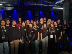 Na scenie w niebieskim świetle stoi duża grupa nastolatków oraz jedna dorosła kobieta. Wszyscy pozują, większość ubrana w czarne koszulki z logo wydarzenia. Tło stanowią ceglane ściany i sprzęt oświetleniowy.