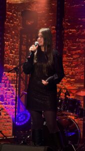 Młoda kobieta w czarnej, błyszczącej sukience śpiewa do mikrofonu. Stoi na tle czerwono-pomarańczowej ceglanej ściany i perkusji.