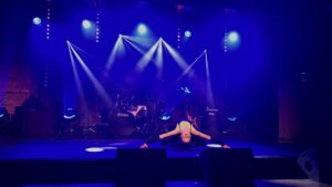 Na scenie w mocnym niebieskim oświetleniu tancerka wykonuje niski, ekspresyjny układ – leży wygięta do tyłu z szeroko rozłożonymi rękami. W tle perkusja i światła sceniczne.