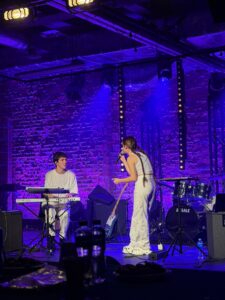 Dziewczyna i chłopak występują razem – chłopak gra na klawiszach, dziewczyna śpiewa i trzyma mikrofon. Oboje w białych strojach. Tło fioletowo podświetlonej ceglanej ściany.