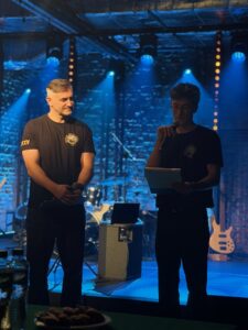 Dwie osoby na scenie: dorosły mężczyzna w czarnej koszulce i nastolatek czytający z kartki do mikrofonu. Za nimi widać instrumenty i niebieskie światła.