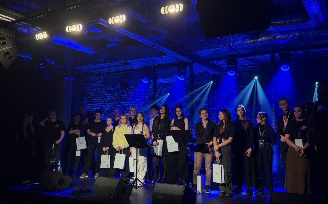 Na scenie w intensywnym niebieskim świetle stoją uczestnicy wydarzenia – kilkanaście osób, część trzyma białe torby z logo. W tle widoczne są reflektory oraz ceglana ściana.