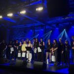 Na scenie w intensywnym niebieskim świetle stoją uczestnicy wydarzenia – kilkanaście osób, część trzyma białe torby z logo. W tle widoczne są reflektory oraz ceglana ściana.