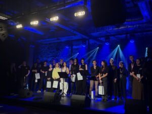 Na scenie w intensywnym niebieskim świetle stoją uczestnicy wydarzenia – kilkanaście osób, część trzyma białe torby z logo. W tle widoczne są reflektory oraz ceglana ściana.