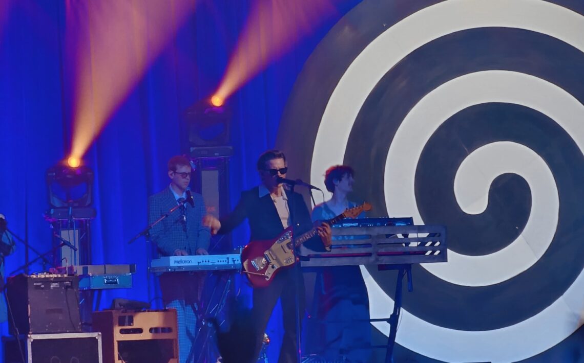 Koncert na scenie z niebieskim tłem i dużą czarno-białą spiralą po prawej stronie. W centrum stoi muzyk w garniturze i okularach przeciwsłonecznych, grający na czerwonej gitarze i śpiewający do mikrofonu. Za nim znajdują się klawiszowcy przy syntezatorach. Nad sceną świecą pomarańczowe reflektory, a na dole widoczne są ciemne sylwetki publiczności.