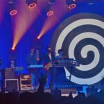 Koncert na scenie z niebieskim tłem i dużą czarno-białą spiralą po prawej stronie. W centrum stoi muzyk w garniturze i okularach przeciwsłonecznych, grający na czerwonej gitarze i śpiewający do mikrofonu. Za nim znajdują się klawiszowcy przy syntezatorach. Nad sceną świecą pomarańczowe reflektory, a na dole widoczne są ciemne sylwetki publiczności.