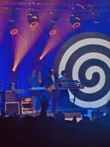 Koncert na scenie z niebieskim tłem i dużą czarno-białą spiralą po prawej stronie. W centrum stoi muzyk w garniturze i okularach przeciwsłonecznych, grający na czerwonej gitarze i śpiewający do mikrofonu. Za nim znajdują się klawiszowcy przy syntezatorach. Nad sceną świecą pomarańczowe reflektory, a na dole widoczne są ciemne sylwetki publiczności.