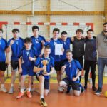 Grupa kilkunastu młodych mężczyzn w niebieskich strojach sportowych pozuje w hali sportowej przed bramką do piłki ręcznej. Część z nich trzyma medale, jeden zawodnik z przodu trzyma złoty puchar, a inny dyplom. Po prawej stronie stoi dorosły mężczyzna, prawdopodobnie trener, również z medalem. Drużyna wygląda na zwycięzców turnieju, wszyscy zwróceni są w stronę aparatu.