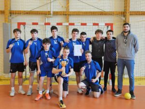 Grupa kilkunastu młodych mężczyzn w niebieskich strojach sportowych pozuje w hali sportowej przed bramką do piłki ręcznej. Część z nich trzyma medale, jeden zawodnik z przodu trzyma złoty puchar, a inny dyplom. Po prawej stronie stoi dorosły mężczyzna, prawdopodobnie trener, również z medalem. Drużyna wygląda na zwycięzców turnieju, wszyscy zwróceni są w stronę aparatu.