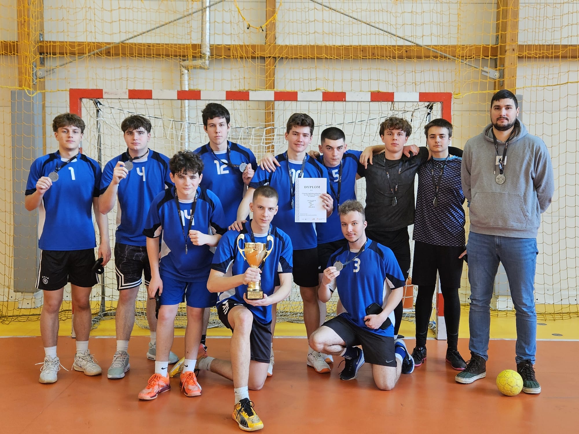 Grupa kilkunastu młodych mężczyzn w niebieskich strojach sportowych pozuje w hali sportowej przed bramką do piłki ręcznej. Część z nich trzyma medale, jeden zawodnik z przodu trzyma złoty puchar, a inny dyplom. Po prawej stronie stoi dorosły mężczyzna, prawdopodobnie trener, również z medalem. Drużyna wygląda na zwycięzców turnieju, wszyscy zwróceni są w stronę aparatu.