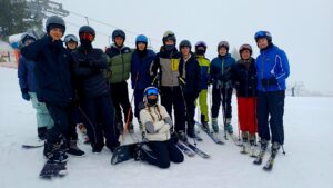 Zbliżone zdjęcie części grupy: młodzież i dorosły instruktor w niebieskiej kurtce stoją na nartach; jedna osoba z snowboardem klęczy na śniegu. Tło stanowi zamglony stok i wyciąg.