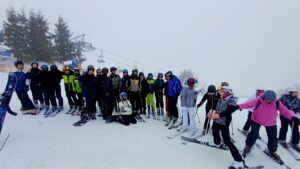 Grupa młodzieży i dorosłych stoi na ośnieżonym stoku narciarskim; wszyscy w kaskach i goglach, część na nartach, część na snowboardach. W tle wyciąg krzesełkowy i drzewa, widoczna mgła.