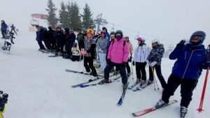 Ujęcie z boku grupy na stoku; osoby stoją na nartach, trzymają kijki, patrzą w stronę aparatu. Widoczny wyciąg narciarski i ośnieżona przestrzeń wokół.