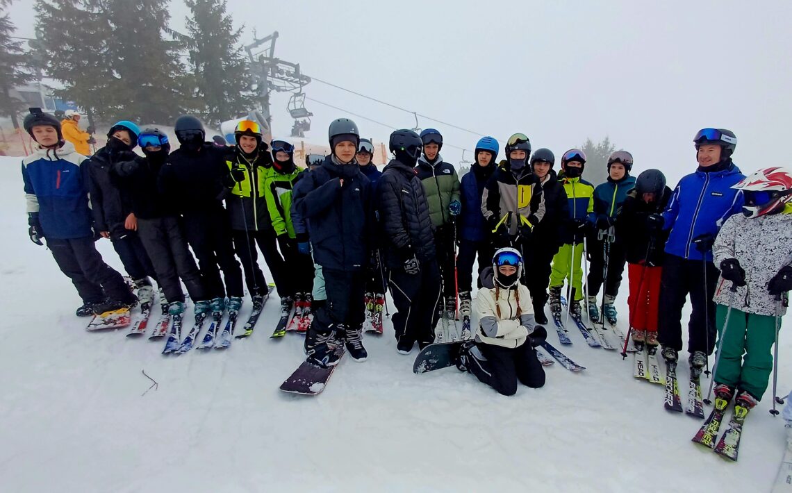 Grupa narciarzy i snowboardzistów pozujących na śniegu; jedna osoba klęczy z przodu, reszta stoi w półokręgu. Za nimi wyciąg krzesełkowy oraz drzewa iglaste w mgle.
