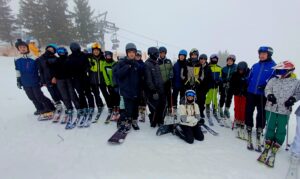 Grupa narciarzy i snowboardzistów pozujących na śniegu; jedna osoba klęczy z przodu, reszta stoi w półokręgu. Za nimi wyciąg krzesełkowy oraz drzewa iglaste w mgle.