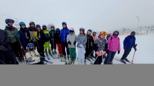 Ta sama grupa ustawiona frontalnie do aparatu na stoku; kolorowe kurtki i kaski wyraźnie kontrastują z białym śniegiem. W tle zamglony stok i elementy infrastruktury narciarskiej.