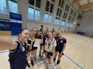 Selfie grupowe wykonane w hali sportowej. Siedem młodych zawodniczek drużyny siatkarskiej stoi blisko siebie na parkiecie, uśmiechnięte i w dobrych nastrojach. Jedna z nich trzyma złoty puchar, inna dyplom, kilka ma medale na szyi. Zawodniczki ubrane są w sportowe stroje (granatowe i białe koszulki, krótkie spodenki, ochraniacze na kolana). W tle widać wysokie okna, siatkę ochronną oraz elementy hali sportowej. Zdjęcie wyraża radość ze zwycięstwa.