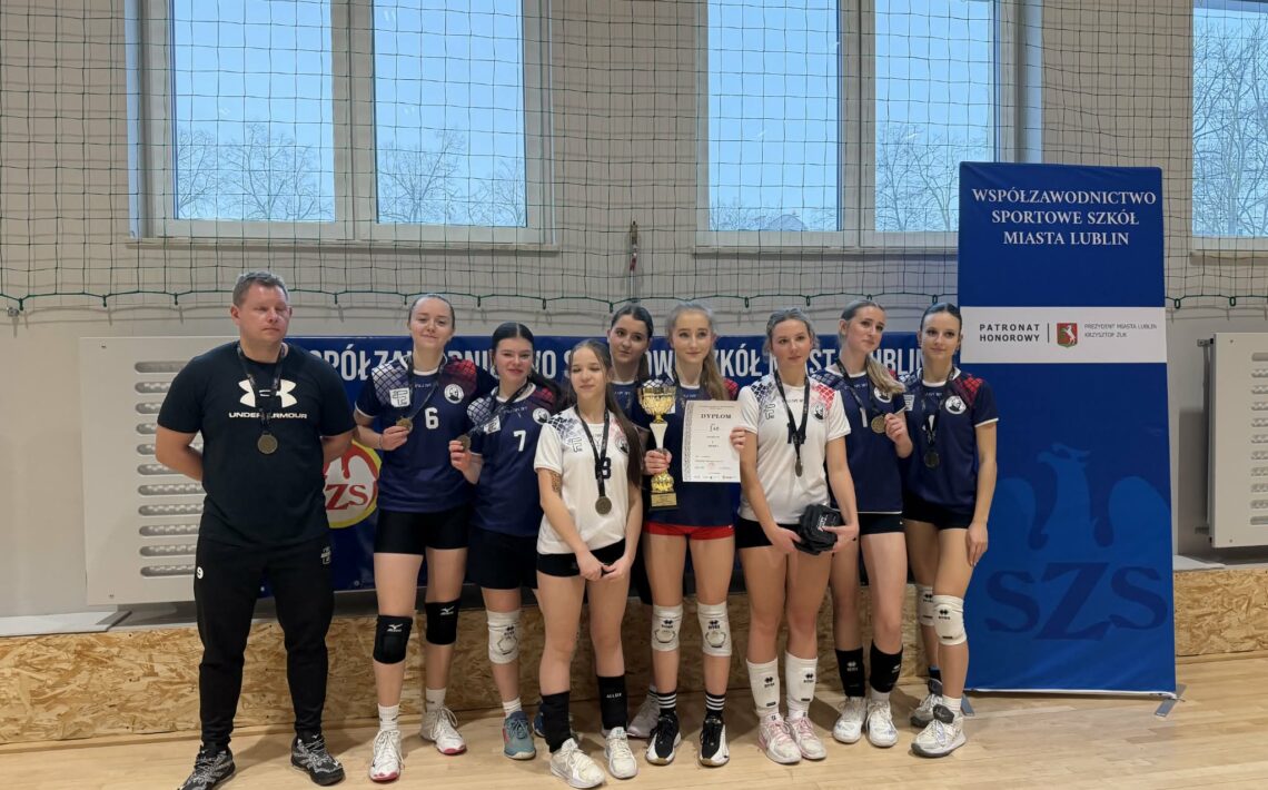 Pozowane zdjęcie całej drużyny siatkarskiej z trenerem w hali sportowej. Dziewięć zawodniczek stoi w jednym rzędzie, obok nich trener ubrany na czarno. Zawodniczki mają na szyjach medale, jedna trzyma puchar, inna dyplom. W tle znajduje się duży baner z czytelnym napisem: „Współzawodnictwo Sportowe Szkół Miasta Lublin” oraz logotypy organizatorów. Zdjęcie dokumentuje oficjalne zakończenie zawodów i zdobycie nagród.