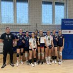 Pozowane zdjęcie całej drużyny siatkarskiej z trenerem w hali sportowej. Dziewięć zawodniczek stoi w jednym rzędzie, obok nich trener ubrany na czarno. Zawodniczki mają na szyjach medale, jedna trzyma puchar, inna dyplom. W tle znajduje się duży baner z czytelnym napisem: „Współzawodnictwo Sportowe Szkół Miasta Lublin” oraz logotypy organizatorów. Zdjęcie dokumentuje oficjalne zakończenie zawodów i zdobycie nagród.