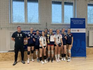Pozowane zdjęcie całej drużyny siatkarskiej z trenerem w hali sportowej. Dziewięć zawodniczek stoi w jednym rzędzie, obok nich trener ubrany na czarno. Zawodniczki mają na szyjach medale, jedna trzyma puchar, inna dyplom. W tle znajduje się duży baner z czytelnym napisem:
„Współzawodnictwo Sportowe Szkół Miasta Lublin” oraz logotypy organizatorów. Zdjęcie dokumentuje oficjalne zakończenie zawodów i zdobycie nagród.