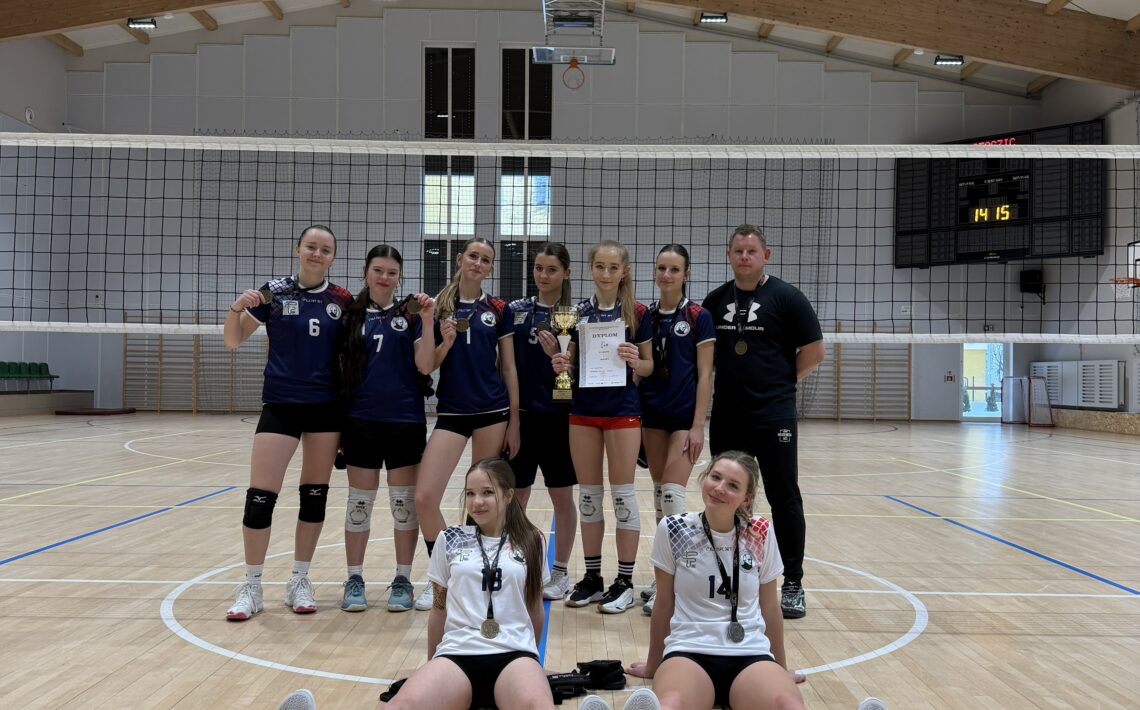 Zdjęcie przedstawia grupę młodych zawodniczek drużyny siatkarskiej w hali sportowej. Dziewięć osób pozuje na środku boiska pod siatką; siedem stoi w tylnej linii, dwie siedzą na podłodze z przodu. Zawodniczki mają na sobie stroje sportowe: większość w granatowych koszulkach i czarnych spodenkach, dwie z przodu w białych koszulkach. Na szyjach widoczne są medale, a jedna z osób trzyma puchar i dyplom, co wskazuje na sukces sportowy. W tle widać drewniany sufit hali, tablicę wyników oraz linie boiska. Atmosfera zdjęcia jest radosna i celebracyjna.