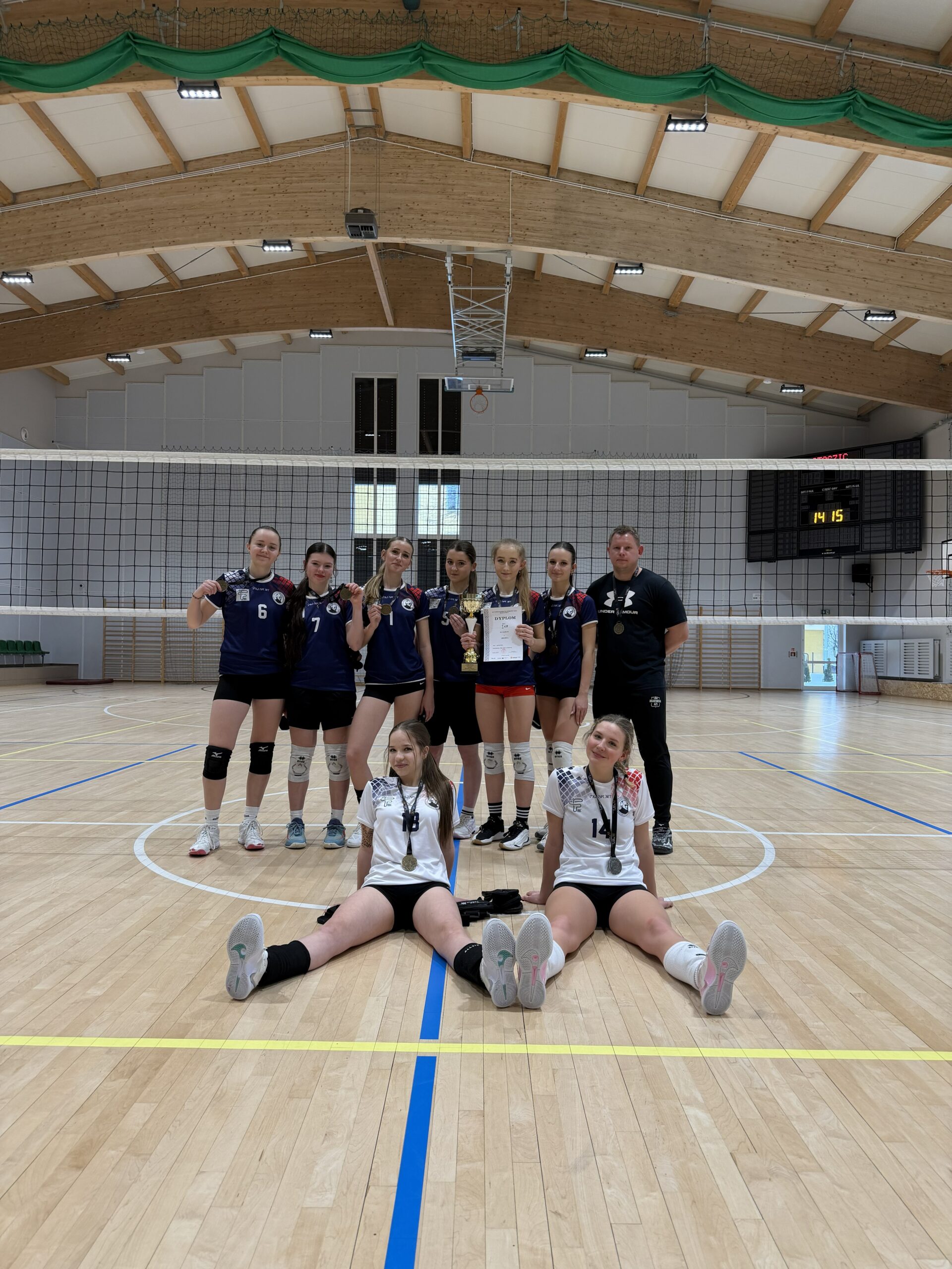 Zdjęcie przedstawia grupę młodych zawodniczek drużyny siatkarskiej w hali sportowej. Dziewięć osób pozuje na środku boiska pod siatką; siedem stoi w tylnej linii, dwie siedzą na podłodze z przodu. Zawodniczki mają na sobie stroje sportowe: większość w granatowych koszulkach i czarnych spodenkach, dwie z przodu w białych koszulkach. Na szyjach widoczne są medale, a jedna z osób trzyma puchar i dyplom, co wskazuje na sukces sportowy. W tle widać drewniany sufit hali, tablicę wyników oraz linie boiska. Atmosfera zdjęcia jest radosna i celebracyjna.