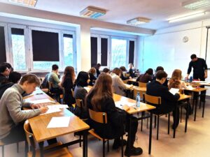 Zdjęcie przedstawia salę lekcyjną podczas pisemnego sprawdzianu lub egzaminu. Kilkanaście uczniów siedzi pojedynczo w ławkach ustawionych w rzędach, wszyscy pochylają się nad kartkami i piszą. Z przodu klasy stoi nauczyciel, który rozdaje lub zbiera arkusze. Pomieszczenie jest jasne, z dużymi oknami po lewej stronie i oświetleniem sufitowym. Na ścianie z przodu widoczna jest tablica i zegar. Atmosfera skupienia i ciszy.