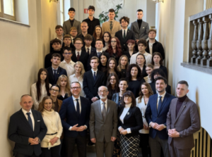 Duża, formalna fotografia grupowa wykonana wewnątrz budynku szkolnego. Na schodach stoi kilkadziesiąt osób: młodzież (uczniowie) oraz dorośli (nauczyciele lub dyrekcja). Wszyscy są ustawieni w kilku rzędach, twarzami do obiektywu. Ubrani są elegancko – dominują garnitury, marynarki, koszule, sukienki i stonowane kolory. Zdjęcie ma charakter oficjalny i pamiątkowy. Tło stanowi jasny korytarz z poręczą schodów i ścianami budynku.