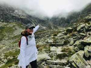 Uśmiechnięta osoba w jasnej kurtce i czapce stoi na kamienistym górskim szlaku, wskazując ręką na mglistą, skalistą dolinę otoczoną stromymi zboczami; w tle surowy krajobraz gór we mgle.