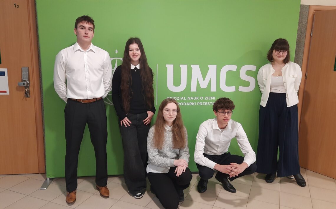 Skrót – Zdjęcie 1 Na zdjęciu widać pięć młodych osób (trzy stojące, dwie w przysiadzie) ustawionych na tle zielonej ścianki z dużym białym napisem „UMCS” oraz mniejszym tekstem „Wydział Nauk o Ziemi i Gospodarki Przestrzennej”. Znajdują się w szkolnym lub akademickim korytarzu – po bokach widoczne są drewniane drzwi z tabliczkami informacyjnymi. Osoby są elegancko ubrane – białe koszule, ciemne spodnie i spódnice. Patrzą w stronę aparatu, przyjmując formalną, pozowaną postawę.