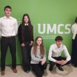 Skrót – Zdjęcie 1 Na zdjęciu widać pięć młodych osób (trzy stojące, dwie w przysiadzie) ustawionych na tle zielonej ścianki z dużym białym napisem „UMCS” oraz mniejszym tekstem „Wydział Nauk o Ziemi i Gospodarki Przestrzennej”. Znajdują się w szkolnym lub akademickim korytarzu – po bokach widoczne są drewniane drzwi z tabliczkami informacyjnymi. Osoby są elegancko ubrane – białe koszule, ciemne spodnie i spódnice. Patrzą w stronę aparatu, przyjmując formalną, pozowaną postawę.