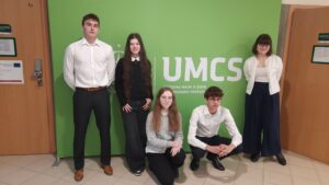 Skrót – Zdjęcie 1 Na zdjęciu widać pięć młodych osób (trzy stojące, dwie w przysiadzie) ustawionych na tle zielonej ścianki z dużym białym napisem „UMCS” oraz mniejszym tekstem „Wydział Nauk o Ziemi i Gospodarki Przestrzennej”. Znajdują się w szkolnym lub akademickim korytarzu – po bokach widoczne są drewniane drzwi z tabliczkami informacyjnymi. Osoby są elegancko ubrane – białe koszule, ciemne spodnie i spódnice. Patrzą w stronę aparatu, przyjmując formalną, pozowaną postawę.