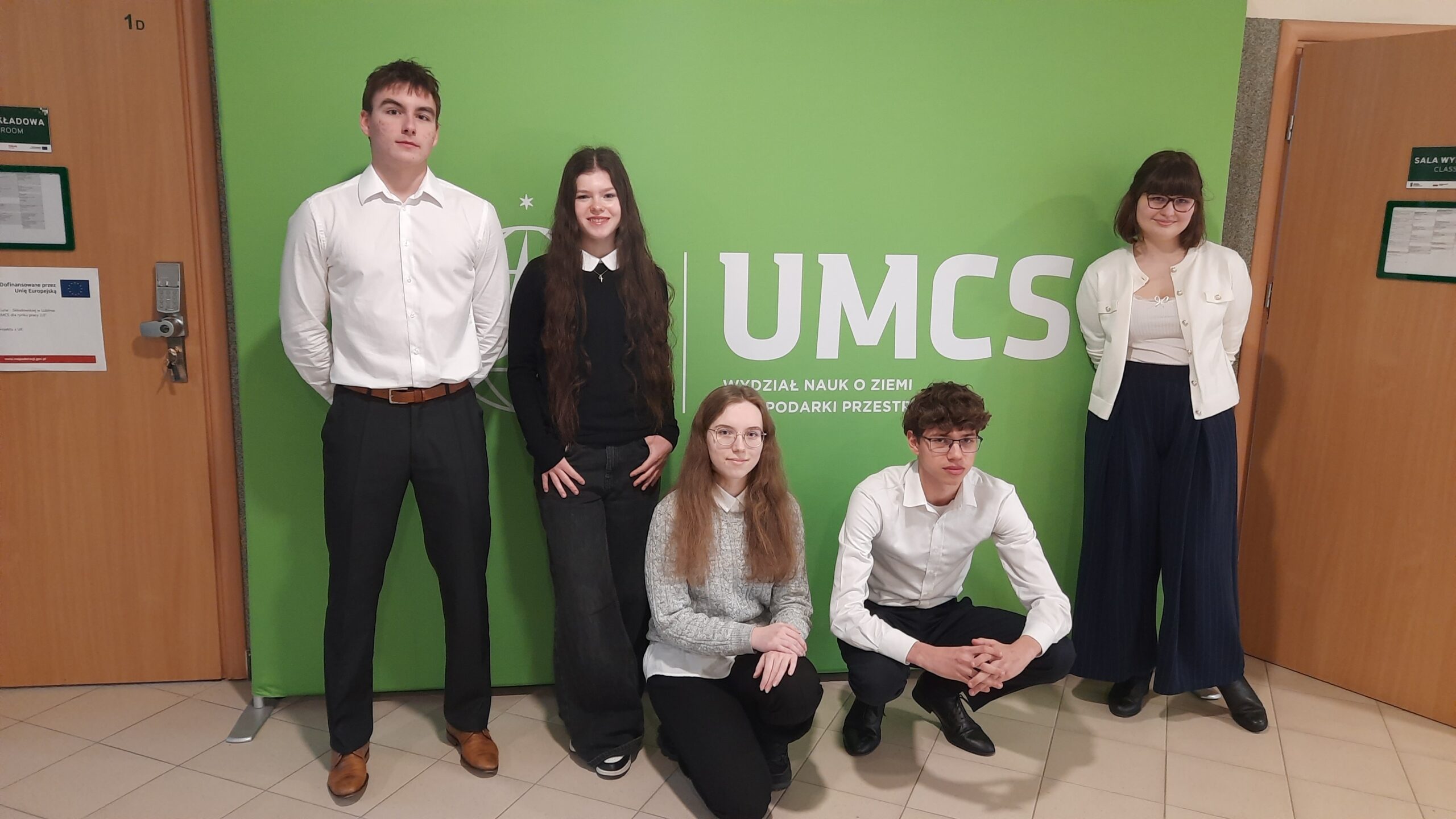 Skrót – Zdjęcie 1 Na zdjęciu widać pięć młodych osób (trzy stojące, dwie w przysiadzie) ustawionych na tle zielonej ścianki z dużym białym napisem „UMCS” oraz mniejszym tekstem „Wydział Nauk o Ziemi i Gospodarki Przestrzennej”. Znajdują się w szkolnym lub akademickim korytarzu – po bokach widoczne są drewniane drzwi z tabliczkami informacyjnymi. Osoby są elegancko ubrane – białe koszule, ciemne spodnie i spódnice. Patrzą w stronę aparatu, przyjmując formalną, pozowaną postawę.