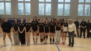 Następna grupa medalistek stojących w szeregu. Wszystkie mają medale na szyjach, część w strojach sportowych, część w odzieży codziennej. Jedna zawodniczka trzyma dyplom i puchar.