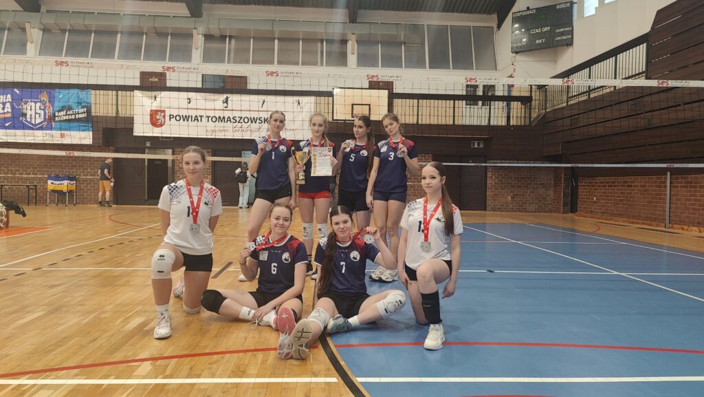 Zdjęcie przedstawia grupę ośmiu młodych zawodniczek drużyny siatkarskiej pozujących w hali sportowej po zakończonych zawodach. Cztery dziewczyny stoją z tyłu, trzy siedzą na parkiecie z przodu, jedna klęczy z boku. Wszystkie mają na szyjach srebrne medale na czerwonych wstążkach. Jedna z zawodniczek trzyma puchar, inna dyplom. Ubrane są w sportowe stroje w kolorze granatowym i białym z numerami. W tle widoczna jest siatka do siatkówki, tablica wyników oraz baner z napisem „Powiat Tomaszowski”. Zdjęcie dokumentuje sukces drużyny w zawodach sportowych.