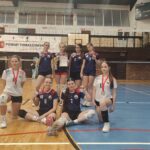 Zdjęcie przedstawia grupę ośmiu młodych zawodniczek drużyny siatkarskiej pozujących w hali sportowej po zakończonych zawodach. Cztery dziewczyny stoją z tyłu, trzy siedzą na parkiecie z przodu, jedna klęczy z boku. Wszystkie mają na szyjach srebrne medale na czerwonych wstążkach. Jedna z zawodniczek trzyma puchar, inna dyplom. Ubrane są w sportowe stroje w kolorze granatowym i białym z numerami. W tle widoczna jest siatka do siatkówki, tablica wyników oraz baner z napisem „Powiat Tomaszowski”. Zdjęcie dokumentuje sukces drużyny w zawodach sportowych.