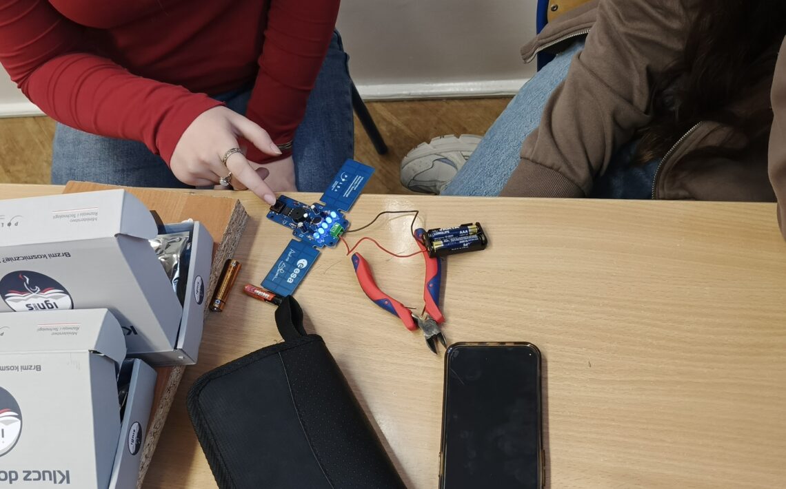 Dwie osoby siedzą przy szkolnej ławce. Na blacie leży uruchomiona niebieska płytka elektroniczna – cztery diody LED świecą jasnym, niebieskim światłem. Do płytki podłączony jest pojemnik na baterie z trzema bateriami AAA, połączony czerwonym i czarnym przewodem. Obok znajdują się czerwono-niebieskie szczypce boczne. Na stole leży również czarny telefon komórkowy ekranem do góry. Po lewej stronie stoją dwa białe pudełka z nadrukiem „IGNIS” i tekstem „Klucz do Kosmosu”. Jedna z osób wskazuje palcem na działającą płytkę, druga obserwuje efekt.