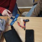 Dwie osoby siedzą przy szkolnej ławce. Na blacie leży uruchomiona niebieska płytka elektroniczna – cztery diody LED świecą jasnym, niebieskim światłem. Do płytki podłączony jest pojemnik na baterie z trzema bateriami AAA, połączony czerwonym i czarnym przewodem. Obok znajdują się czerwono-niebieskie szczypce boczne. Na stole leży również czarny telefon komórkowy ekranem do góry. Po lewej stronie stoją dwa białe pudełka z nadrukiem „IGNIS” i tekstem „Klucz do Kosmosu”. Jedna z osób wskazuje palcem na działającą płytkę, druga obserwuje efekt.