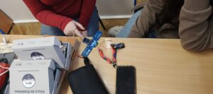 Dwie osoby siedzą przy szkolnej ławce. Na blacie leży uruchomiona niebieska płytka elektroniczna – cztery diody LED świecą jasnym, niebieskim światłem. Do płytki podłączony jest pojemnik na baterie z trzema bateriami AAA, połączony czerwonym i czarnym przewodem. Obok znajdują się czerwono-niebieskie szczypce boczne. Na stole leży również czarny telefon komórkowy ekranem do góry. Po lewej stronie stoją dwa białe pudełka z nadrukiem „IGNIS” i tekstem „Klucz do Kosmosu”. Jedna z osób wskazuje palcem na działającą płytkę, druga obserwuje efekt.