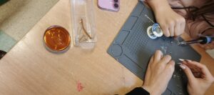 Na drewnianym blacie znajduje się szara mata silikonowa do lutowania z przegródkami. Dwie osoby pracują nad drobnymi elementami elektronicznymi – widać ich dłonie. Jedna osoba trzyma lutownicę, druga cienki element (prawdopodobnie diodę LED lub rezystor). Na macie leżą kawałki cyny. Po lewej stronie stoi okrągłe metalowe pudełko z bursztynową kalafonią. Obok znajduje się przezroczyste plastikowe pudełko z odcinkami przewodów. Na blacie widać także małe różowe fragmenty izolacji przewodów.