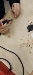 Na jasnym blacie biurka widoczne są dłonie osoby lutującej element elektroniczny. W prawej dłoni trzyma lutownicę z czarno-czerwoną rączką i metalowym grotem, który dotyka wygiętego przewodu. W lewej dłoni trzymany jest cienki drucik. Na palcu widoczny jest pierścionek z dużym, jasnym kamieniem. Na blacie leżą małe elementy elektroniczne: rezystory z paskami, diody, krótkie odcinki drutu oraz okrągły czarny brzęczyk z przewodami. Widać drobne ślady kalafonii i cyny. W dolnej części zdjęcia przebiega czarny kabel zasilający lutownicę.