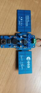 Na jasnym, drewnianym blacie leży niebieska płytka drukowana (moduł elektroniczny) o prostokątnym kształcie z zaokrąglonymi narożnikami. Po prawej stronie znajduje się zielone złącze z oznaczeniami „+” i „–”, do którego podłączone są czerwony i czarny przewód. Obok widoczne są cztery przezroczyste, niebieskie diody LED ustawione w pionowym rzędzie. Na płytce znajdują się również: czarny okrągły brzęczyk (z małym otworem pośrodku), mikroprzycisk, układ scalony w czarnej obudowie, kilka rezystorów z kolorowymi paskami, kondensatory oraz tranzystory. Po obu stronach płytki umieszczono niebieskie prostokątne panele z nadrukiem. Na górnym widnieje tekst: „Ministerstwo Rozwoju i Technologii” oraz „POLSA Polska Agencja Kosmiczna”. Na dolnym panelu znajduje się logo i napis „esa” oraz odręczny podpis „Astro Sławosz”.