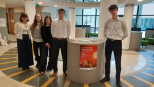 Na zdjęciu widać tę samą grupę pięciu młodych osób stojących w przestronnym holu budynku uczelni. W centrum znajduje się okrągły filar z czerwono-pomarańczowym plakatem „LII Olimpiada Geograficzna”. W tle widoczne są duże okna, białe kolumny i miejsca do siedzenia. Uczestnicy są elegancko ubrani i stoją w luźnym półkolu wokół filaru, patrząc w stronę aparatu. Atmosfera jest oficjalna i związana z wydarzeniem edukacyjnym.