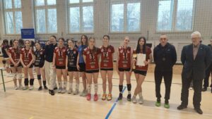 Jedna z drużyn ustawiona w szeregu. Zawodniczki w czerwonych koszulkach, jedna trzyma puchar i dyplom. Obok stoją dorośli opiekunowie. Wszyscy pozują do zdjęcia w hali sportowej.